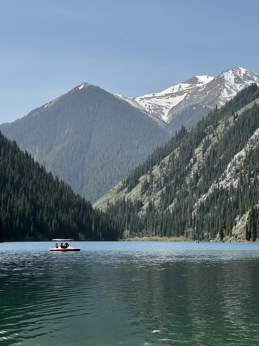 Small pedal boat with passengers on Kolsai Lake, snow-capped peaks rising behind pine-covered slopes